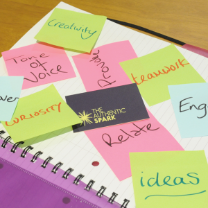 An open notebook on a desk. Blue, pink and green post-it notes are on it with the words 'ideas, engage, relate, provoke, tone of voice, creativity, teamwork,